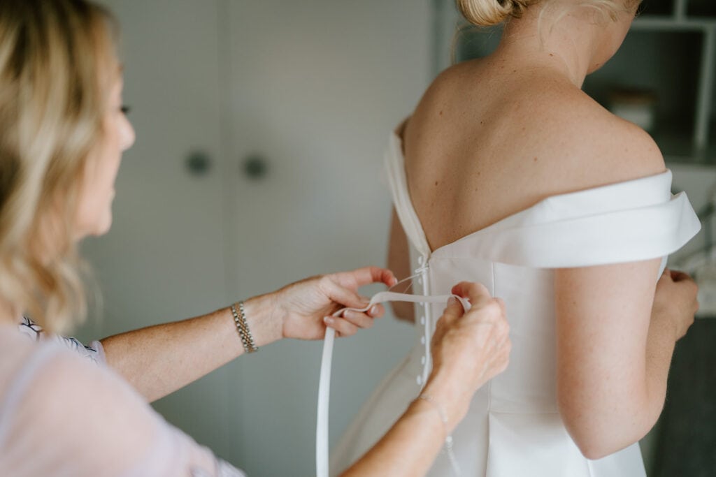 A woman assists another into an off-shoulder white wedding dress at a charming East Quay venue, tying the back with a satin ribbon. The focus on the hands highlights both the gown's elegance and this intimate, preparatory wedding moment. Image by Pearce Wedding Photography.