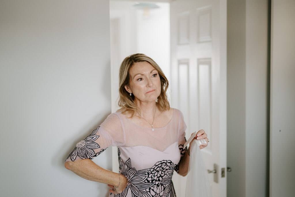 A woman with shoulder-length hair stands in a doorway at the East Quay venue, wearing a dress with floral patterns. She holds a piece of fabric and gazes thoughtfully into the distance, envisioning a wedding. The room is softly lit, creating a calm atmosphere. Image by Pearce Wedding Photography.