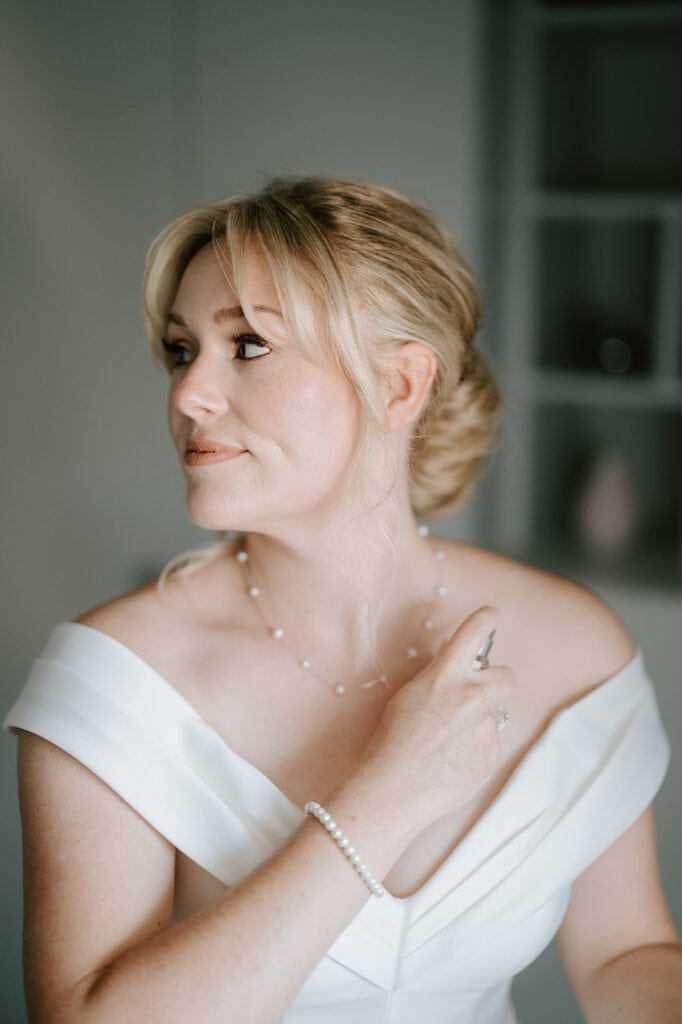 A person in an off-shoulder white dress sprays perfume, with hair styled elegantly in an updo, likely preparing for a wedding at the East Quay Venue. The person wears a pearl necklace and bracelet and is gazing to the side in a softly lit room. Image by Pearce Wedding Photography.