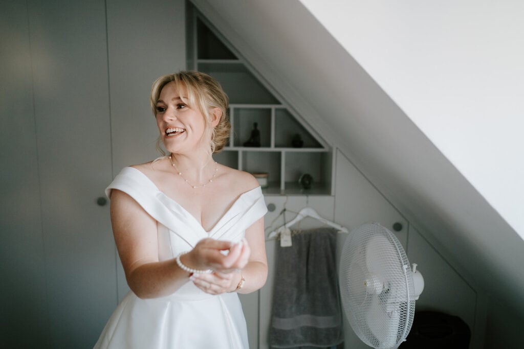 A woman in a white dress smiles while standing in a room with slanted ceilings, reminiscent of the serene atmosphere at the East Quay Venue. Behind her, there's a white fan, shelves lined with small items, and a hanging towel, adding to the calm and intimate setting. Image by Pearce Wedding Photography.