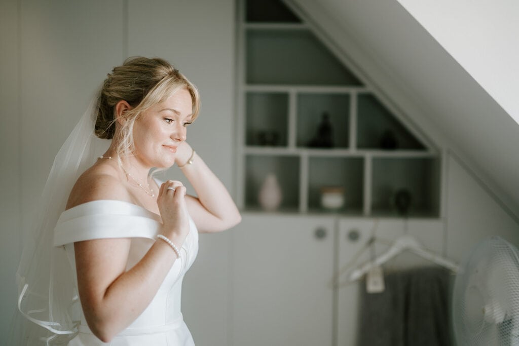 A bride at East Quay Venue, wearing an off-shoulder white dress and veil, stands in a softly lit room, adjusting her earring. Behind her is a built-in shelf with decor items and a wardrobe, all reflecting the gentle, calm expression on her face as she prepares for the wedding. Image by Pearce Wedding Photography.