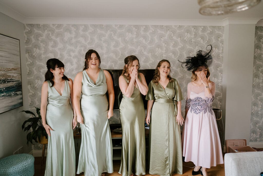 Five women in elegant pastel dresses gather joyfully at the East Quay Venue, surrounded by floral wallpaper. Four wear green dresses, while one, wearing a pink dress and a large black hat, stands out. Their expressions reveal surprise or laughter, capturing the essence of wedding bliss. Image by Pearce Wedding Photography.