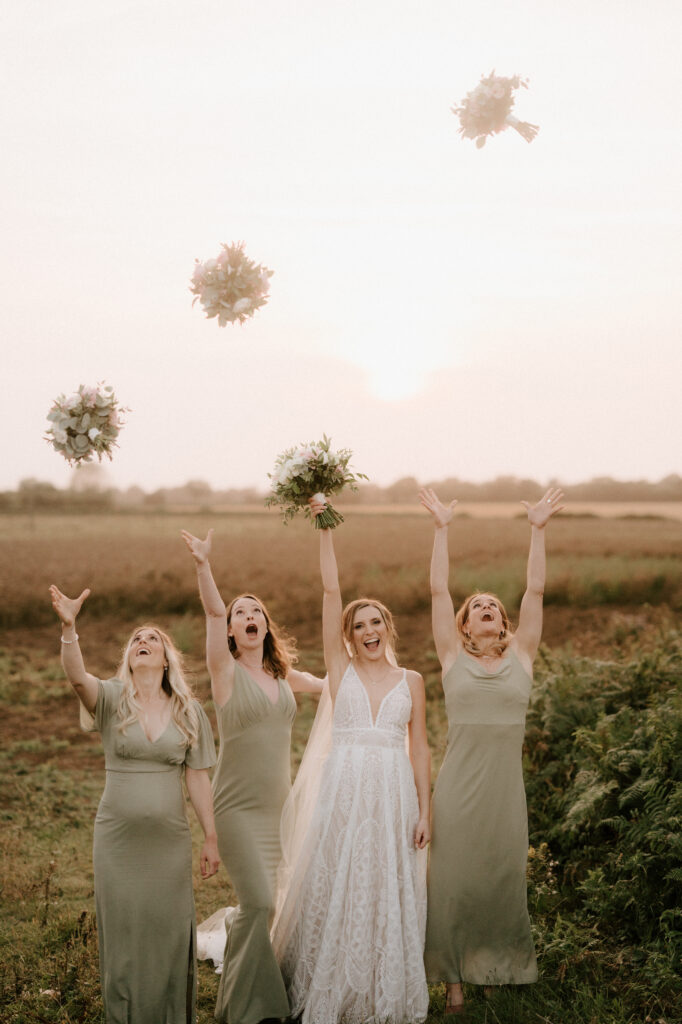 A bride in a white lace gown stands with three bridesmaids in light green dresses, all joyfully throwing their bouquets into the air against the serene sunset backdrop of The Old Kent Barn during golden hour. The women are smiling and appear to be celebrating a beautiful wedding moment. Image by Pearce Wedding Photography.