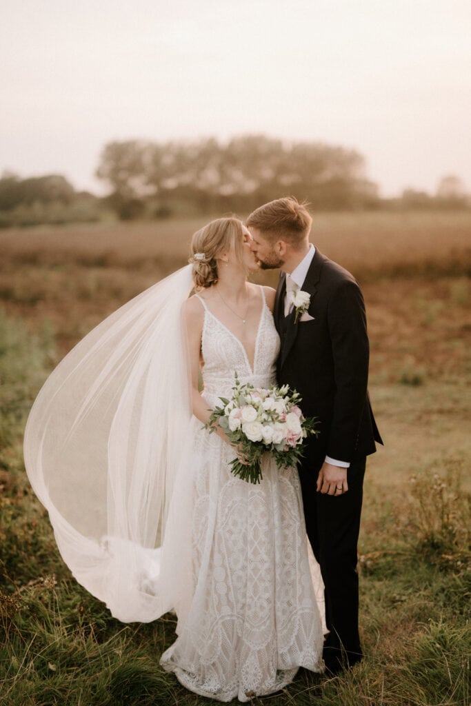 A couple on their wedding day stands in a field at The Old Kent Barn. The bride, in an elegant white gown with a long veil flowing in the wind, holds a bouquet of flowers. The groom, in a black suit, leans in to kiss her forehead as bridesmaids look on. The background is a serene landscape at sunset. Image by Pearce Wedding Photography.