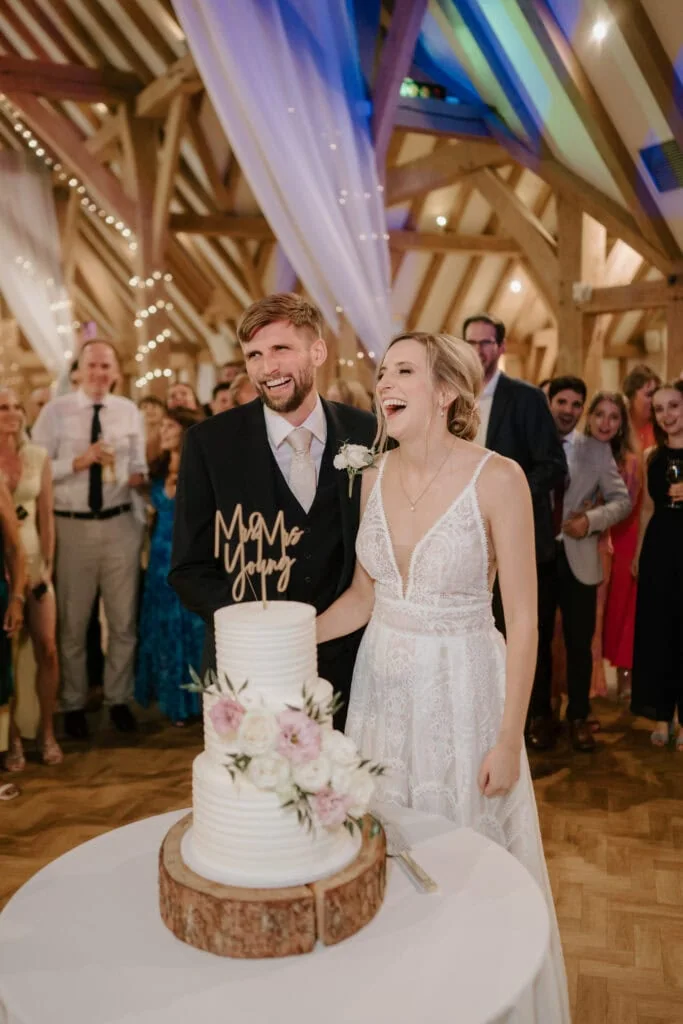 A bride and groom laugh joyfully as they stand beside a tiered wedding cake adorned with pink roses and a "Mr & Mrs" topper. Captured by a Kent wedding photographer, guests in formal attire gather around them in the warmly lit venue. Image by Pearce Wedding Photography.