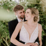 A couple, dressed in wedding attire, stands surrounded by lush, green foliage with small lights in the background at The Old Kent Barn. The groom, with short hair and a beard, embraces the bride from behind. The bride, in a lace wedding dress, smiles as she looks up at him while bridesmaids look on. Image by Pearce Wedding Photography.