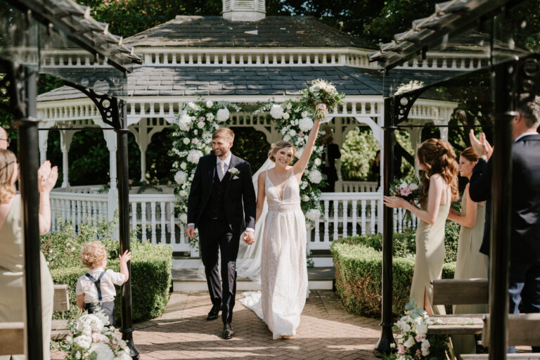 A newlywed couple walks down the aisle outdoors during their enchanting outdoor wedding in Kent, holding hands and smiling. The bride raises her bouquet triumphantly. They are surrounded by greenery and white flowers, with a charming gazebo in the background. Guests on either side applaud and celebrate. Image by Pearce Wedding Photography.