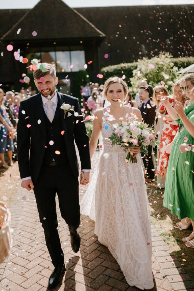 A newlywed couple, dressed in a dark suit and white lace wedding gown, walk hand-in-hand outside The Old Kent Barn as guests shower them with colorful confetti. The bride holds a bouquet of flowers and smiles brightly. The atmosphere is joyful and celebratory, embodying the essence of a perfect wedding day. Image by Pearce Wedding Photography.