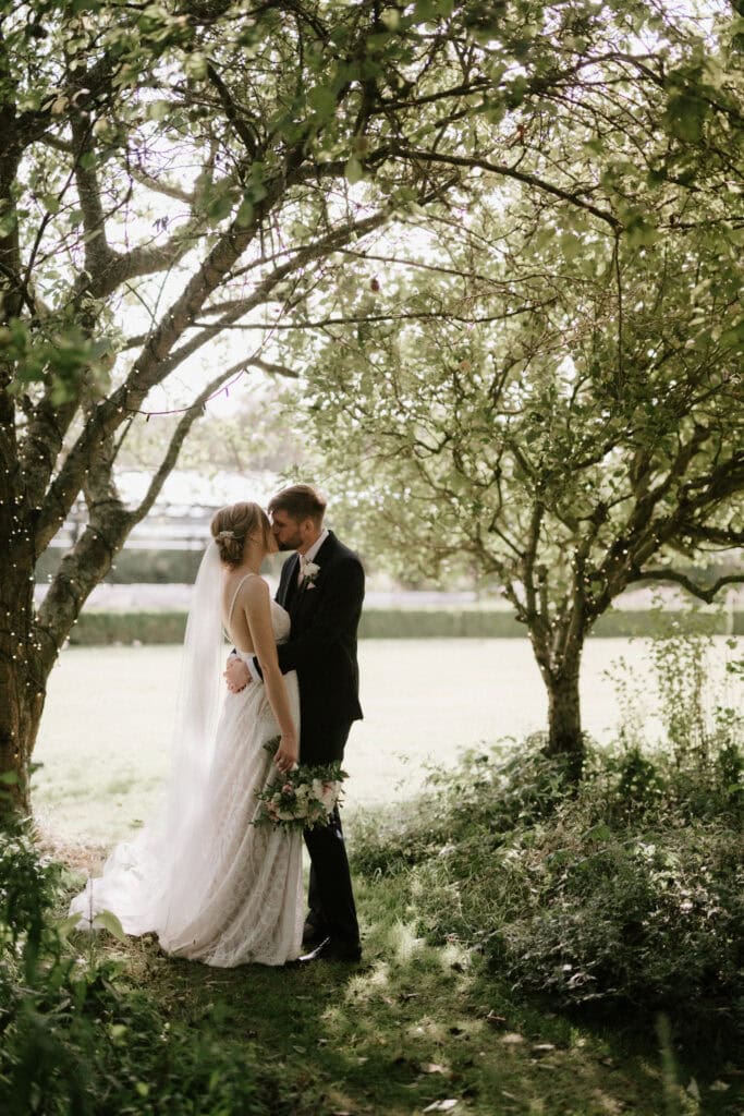 A bride and groom share an intimate moment under a canopy of trees in the lush garden of The Old Kent Barn. The bride, in her white dress and veil, holds a bouquet while the groom stands beside her in a black suit. Sunlight filters through the leaves, enhancing the romantic atmosphere of their wedding day. Image by Pearce Wedding Photography.