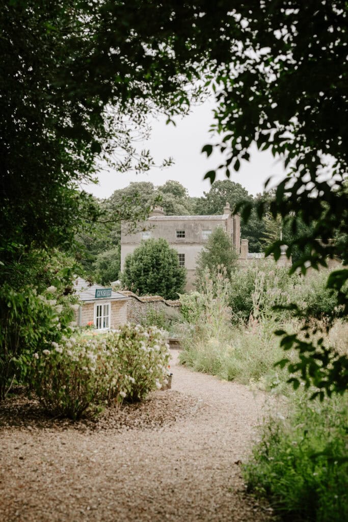 A rustic, two-story stone house partially hidden by lush greenery sits within the charm of Ripple Court Estate. The foreground features a gravel path bordered by shrubs and trees, while the background shows a cloudy sky over a dense forest, creating an idyllic setting perfect for a Deal wedding. Image by Pearce Wedding Photography.