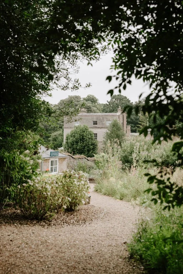 A rustic, two-story stone house partially hidden by lush greenery sits within the charm of Ripple Court Estate. The foreground features a gravel path bordered by shrubs and trees, while the background shows a cloudy sky over a dense forest, creating an idyllic setting perfect for a Deal wedding. Image by Pearce Wedding Photography.