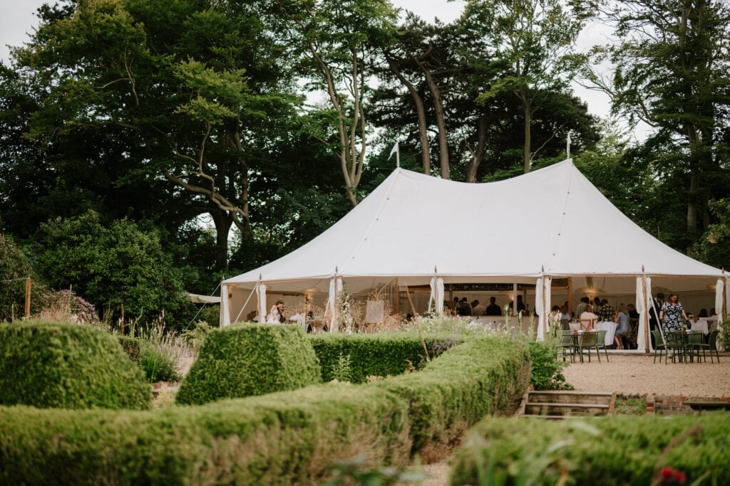 A large white marquee tent set up outdoors at Ripple Court Estate is surrounded by lush greenery and neatly trimmed hedges. People are gathered inside the tent, visible between the tent poles. Tall trees provide a scenic backdrop under a clouded sky during this elegant wedding affair. Image by Pearce Wedding Photography.