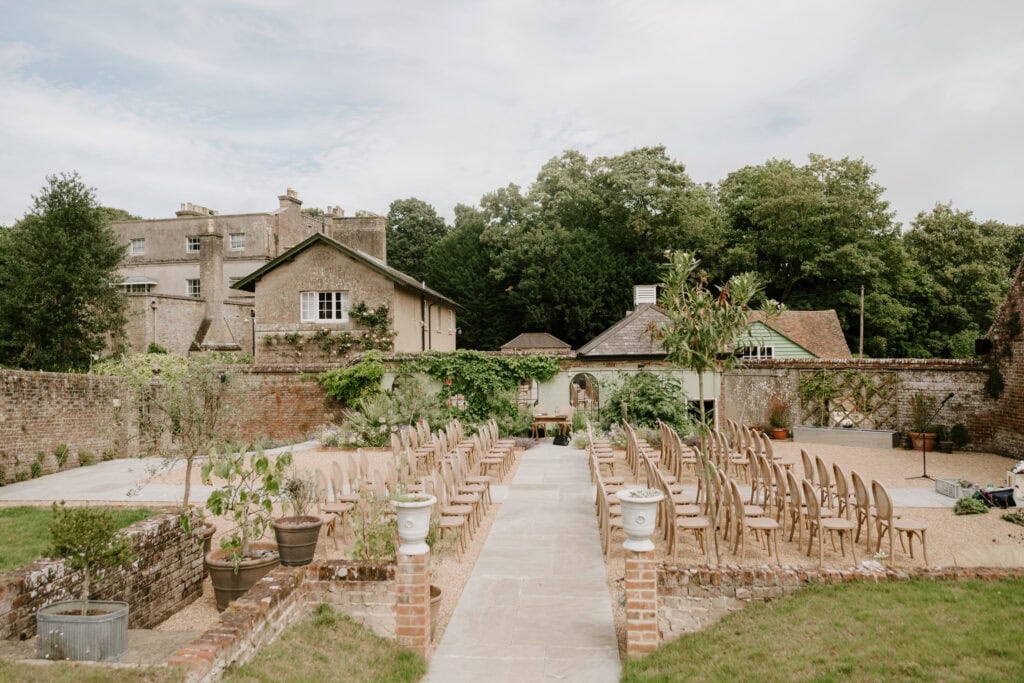An outdoor wedding ceremony setup at Ripple Court Estate features rows of wooden chairs arranged in an aisle formation. Surrounded by greenery, brick buildings, and trees, the venue exudes tranquility. The clear and bright weather adds to the serene atmosphere. Image by Pearce Wedding Photography.