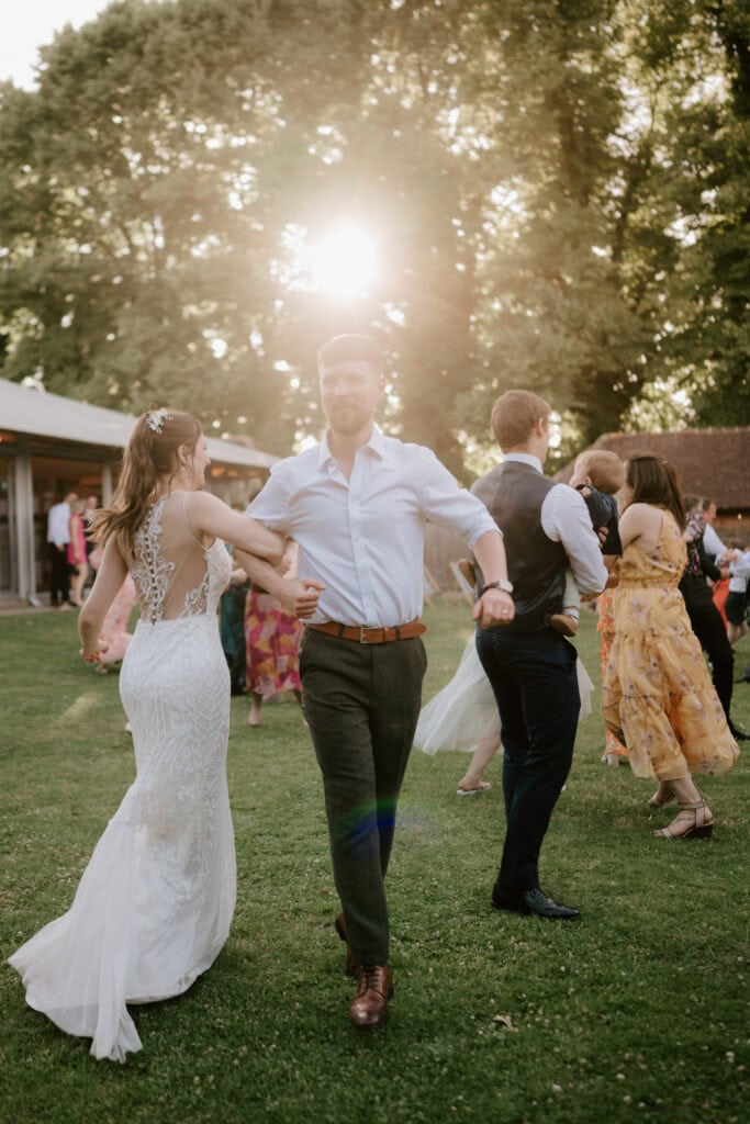 A couple dances outdoors on a sunny day; the woman wears a white wedding dress, and the man is in a white shirt and dark pants. Other couples and guests dance and socialize in the background. Large trees and a building are visible, with sunlight streaming through the leaves, captured by a Canterbury wedding photographer. Image by Pearce Wedding Photography.