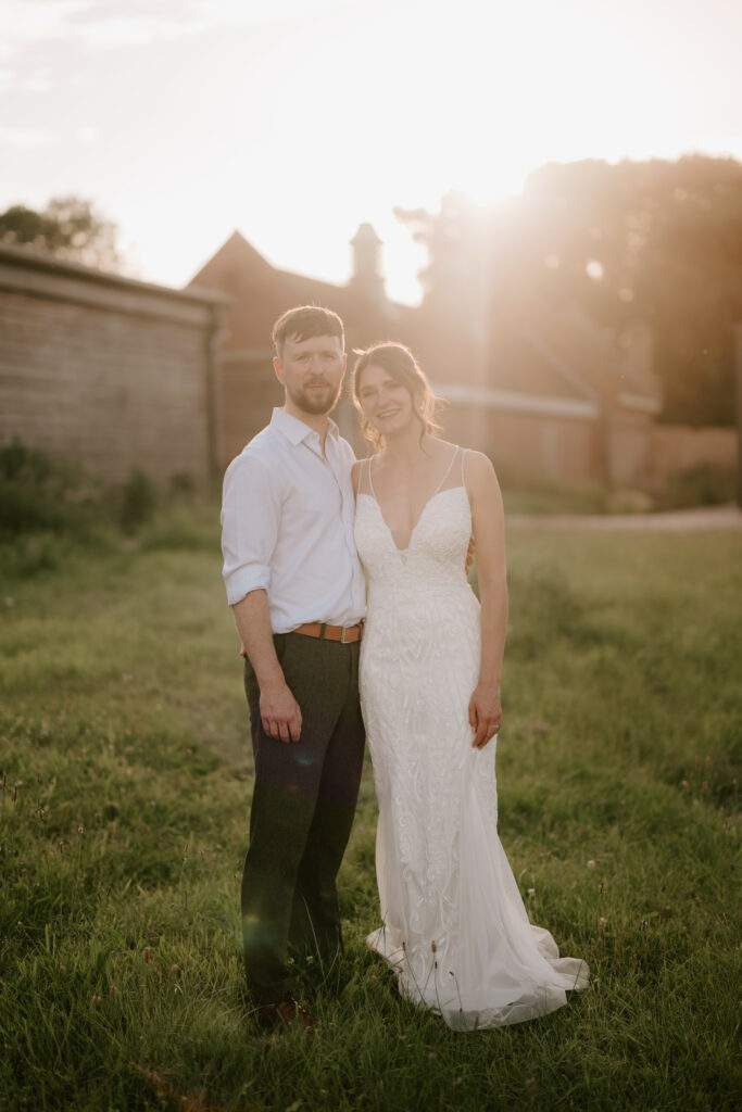 A couple stands together in a grassy field with sunlight shining behind them. The woman is wearing a white, sleeveless wedding dress, and the man is dressed in a white shirt and dark pants. Both are smiling, captured beautifully by their Canterbury wedding photographer. Rustic buildings frame this enchanting moment. Image by Pearce Wedding Photography.