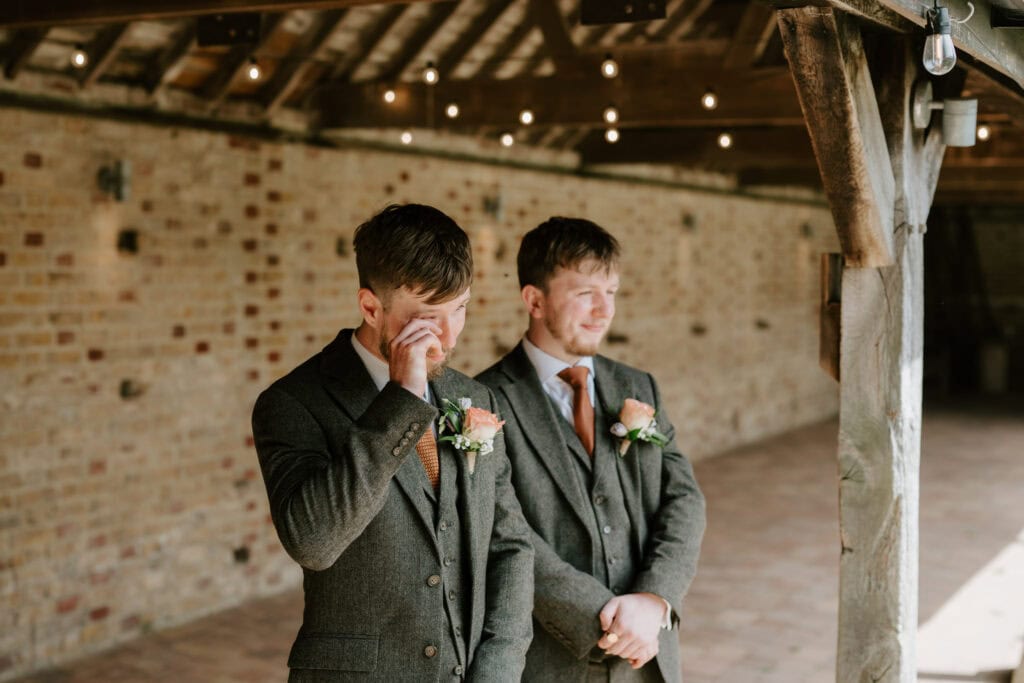 A man in a suit wipes away a tear while another man, also in a suit, stands beside him smiling. They are both wearing boutonnières and standing in the night yard of Kent's rustic wedding venue, with brick walls and string lights above. Image by Pearce Wedding Photography.