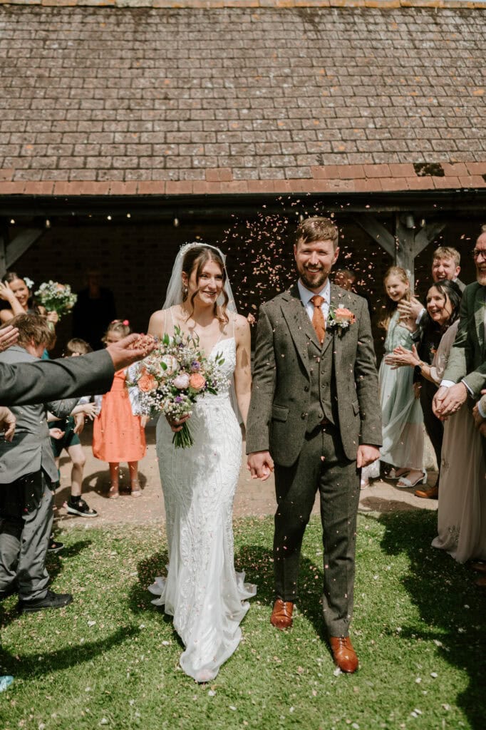 A bride and groom holding hands walk down an outdoor aisle in the night yard while guests toss confetti. The bride wears a white lace dress and holds a bouquet of flowers, and the groom is dressed in a brown suit with a coordinating boutonniere. Smiling friends and family surround them at this enchanting wedding. Image by Pearce Wedding Photography.