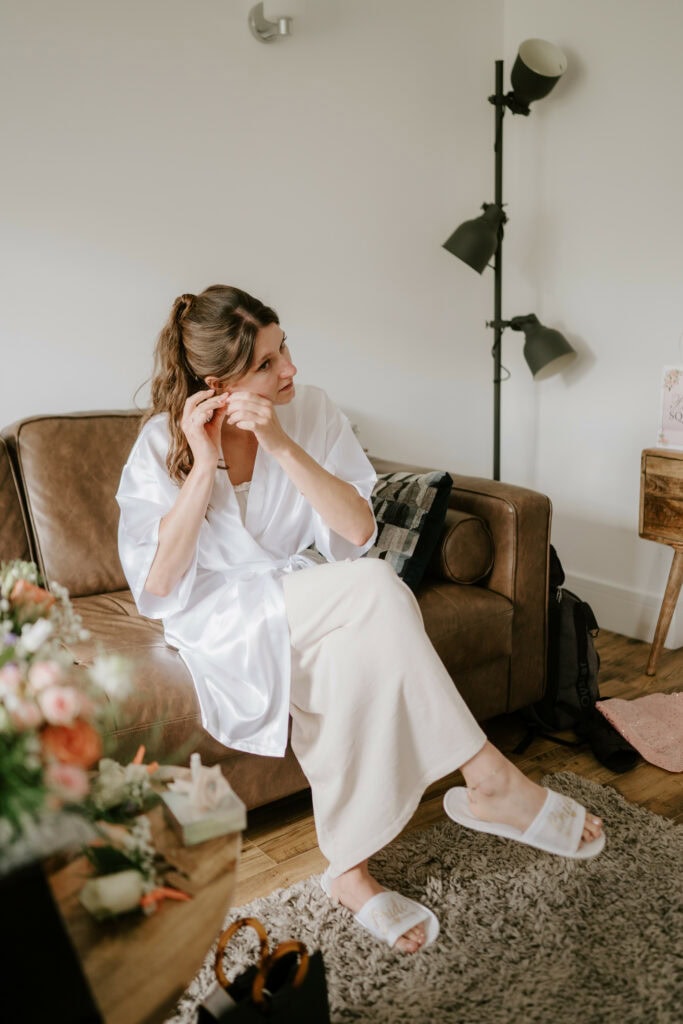 Woman sits adjusting earring, white robe and slippers.