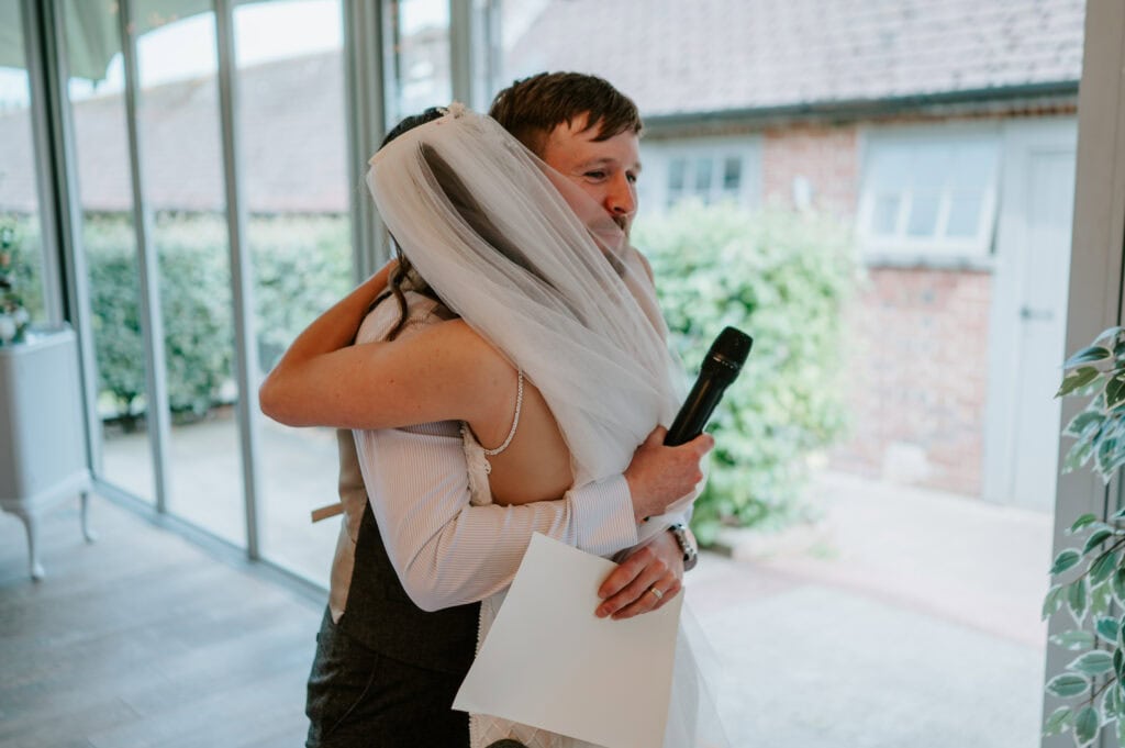 Bride hugs groom holding microphone and paper.