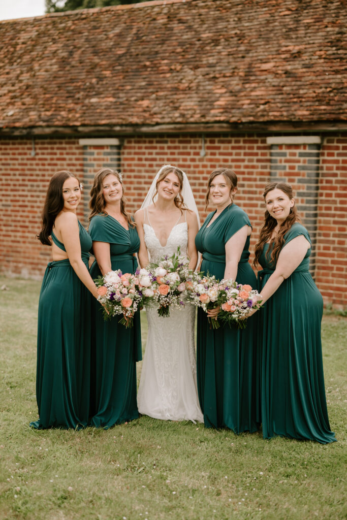 A bride in a white dress stands in the center of four bridesmaids in matching dark green dresses, each holding bouquets of colorful flowers. They are outdoors, posing in front of a brick building with a tiled roof, captured beautifully by the Canterbury wedding photographer. Image by Pearce Wedding Photography.