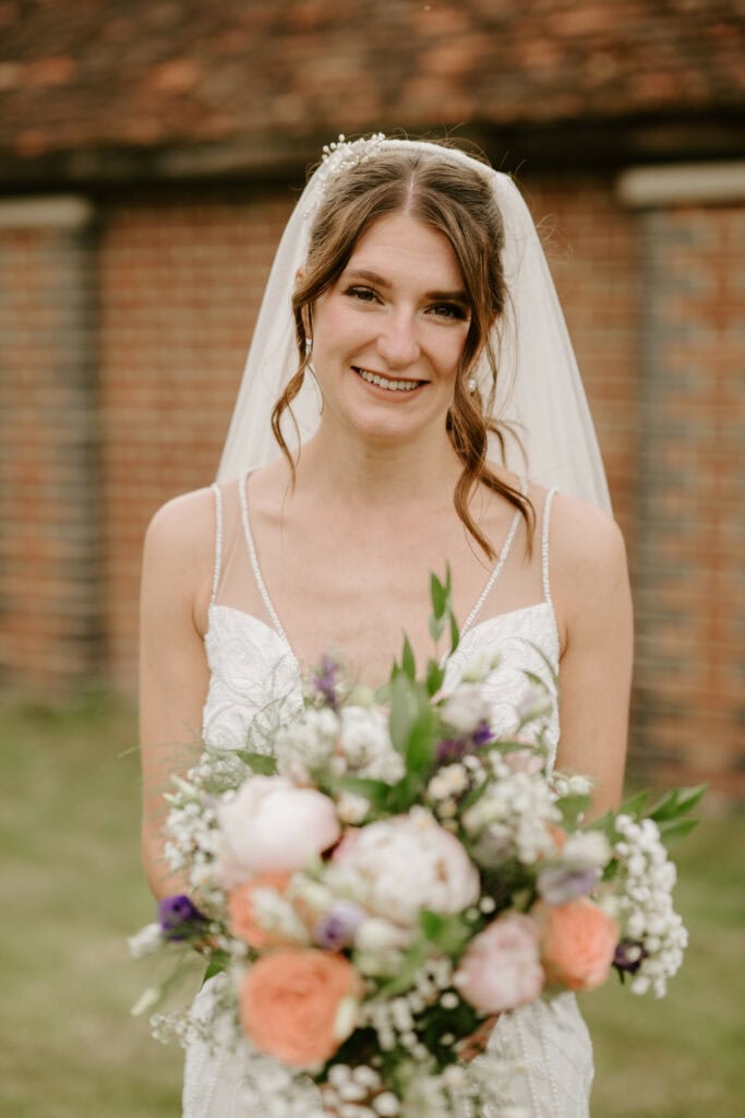 A bride with curled hair and a white veil smiles while holding a bouquet of pink and white blooms. She is wearing a white lace wedding dress and is standing outside in front of a brick wall, capturing the timeless elegance of a Kent wedding. Image by Pearce Wedding Photography.