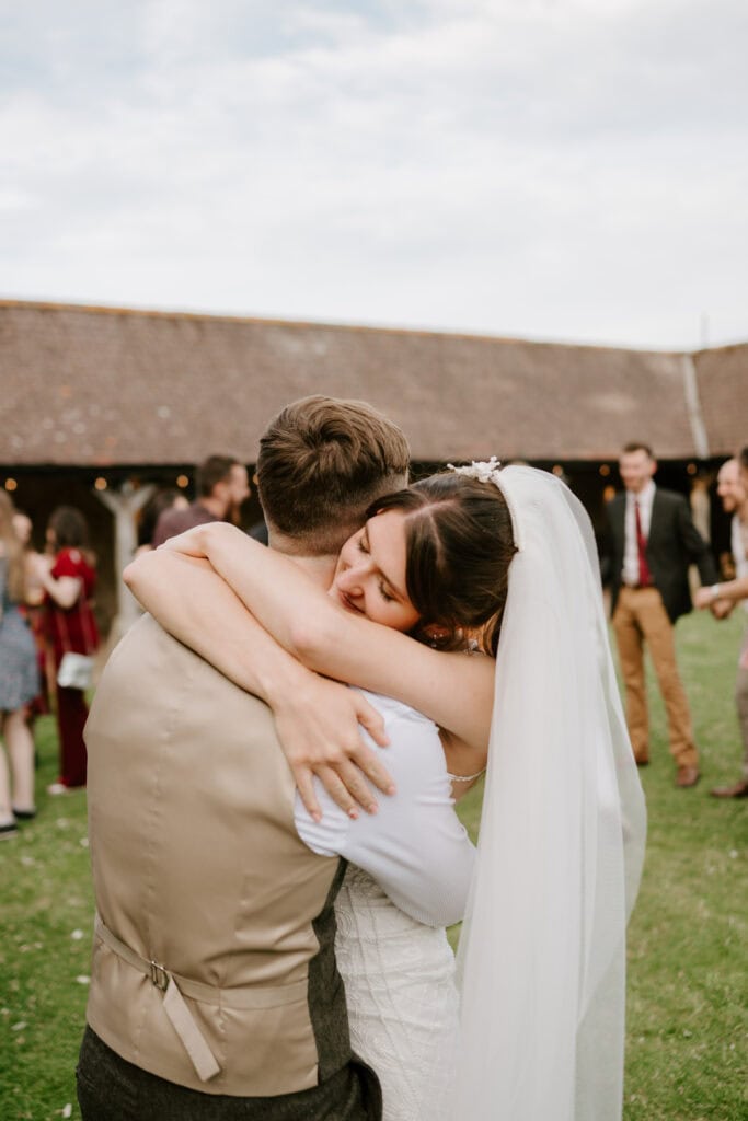 A bride and groom embrace each other lovingly outdoors on a grassy area. The bride, wearing a white dress and veil, has her arms around the groom's neck. The groom, dressed in a vest and shirt, holds her tightly. Their moment is beautifully captured by a Canterbury wedding photographer with guests in the background. Image by Pearce Wedding Photography.