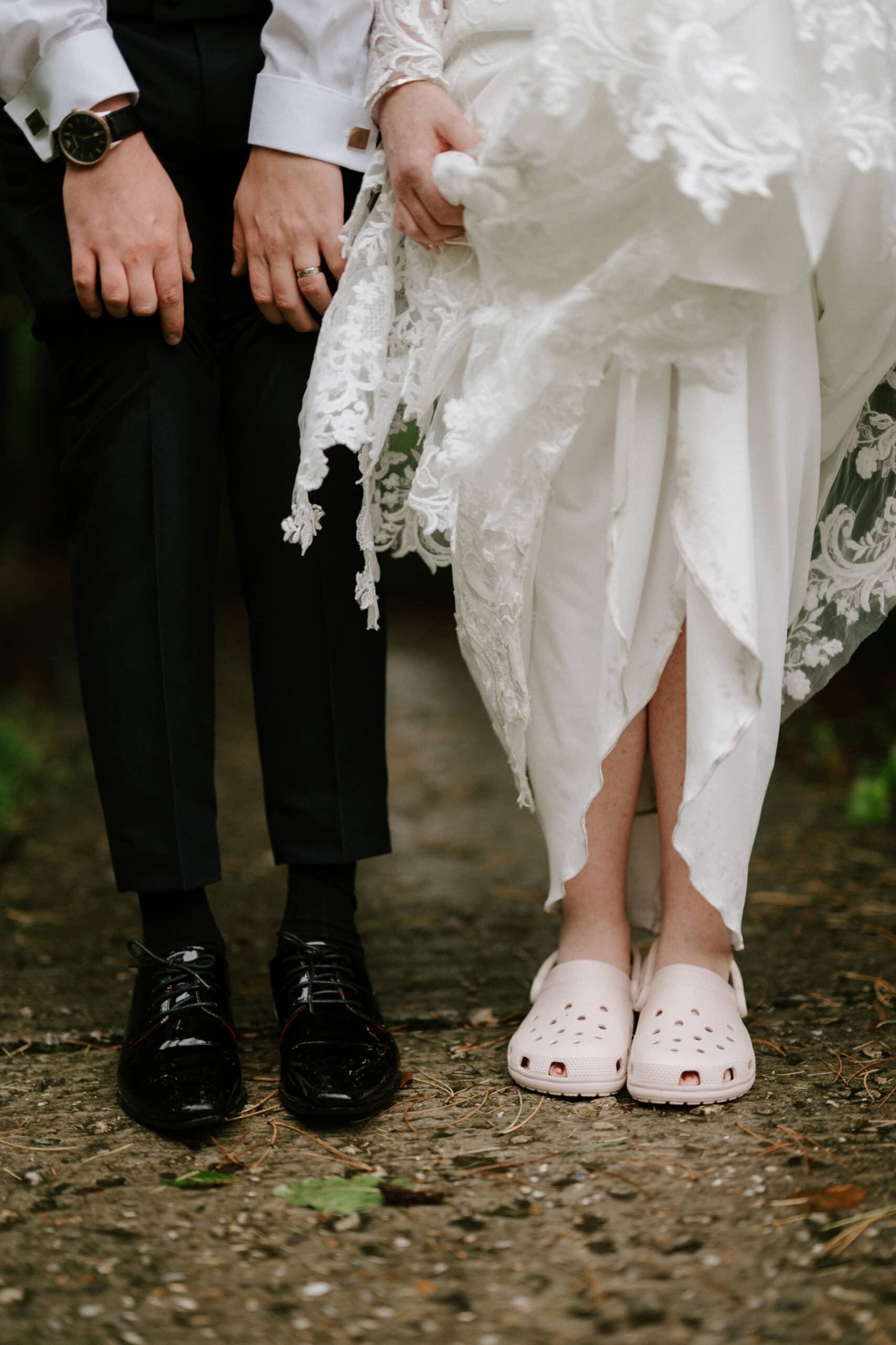A couple stands side by side in wedding attire. The groom wears black formal shoes and a suit, while the bride lifts her lace-trimmed dress slightly, revealing pink Crocs on her feet. The setting is an outdoor path with greenery, creating a whimsical twist to their special day. Image by Pearce Wedding Photography.