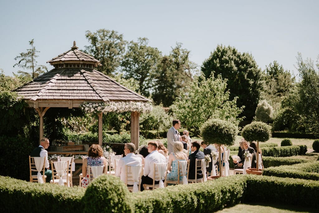 A group of people is seated around tables under a wooden gazebo, surrounded by manicured hedges and lush greenery on a sunny day. The scene, reminiscent of The Secret Garden, suggests a social gathering or outdoor event in a picturesque garden setting. Image by Pearce Wedding Photography.