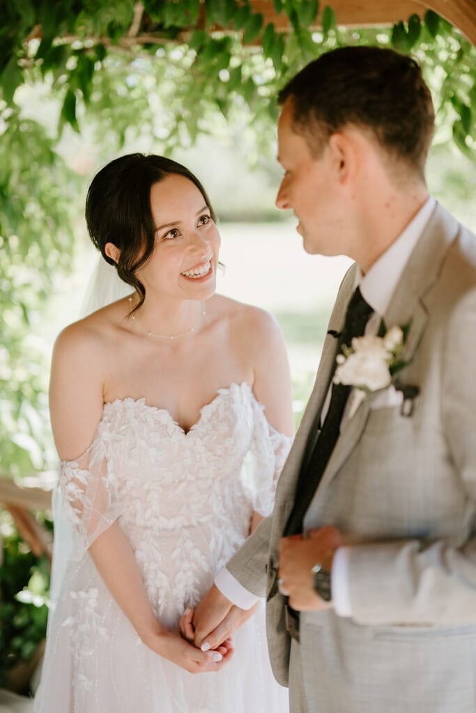 A bride and groom share a joyful moment outdoors under a leafy arbor. The bride wears an off-shoulder white lace wedding dress with a veil, while the groom is in a light gray suit with a boutonniere. Captured by their Ashford Wedding Photographer, they hold hands and gaze into each other’s eyes, smiling. Image by Pearce Wedding Photography.
