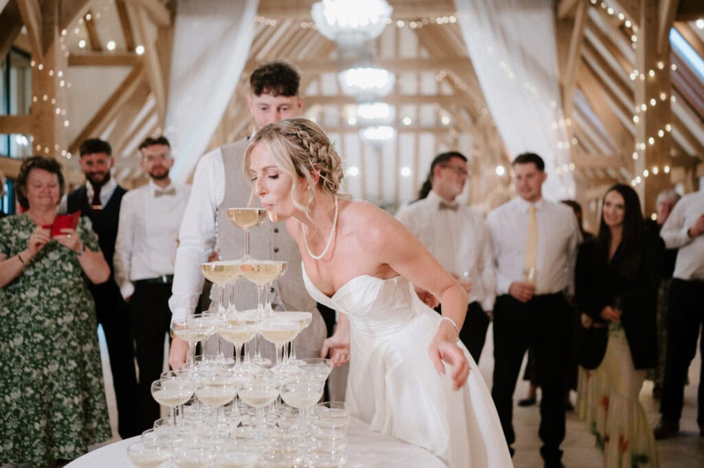 A bride in a white dress leans forward to sip champagne from the top of a tower of glasses at a Kent wedding reception. A wedding photographer snaps the perfect shot as guests stand around, watching and smiling amidst fairy lights and rustic wooden beams. Image by Pearce Wedding Photography.