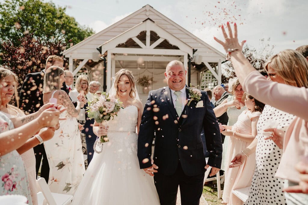A joyous bride and groom walk down the aisle after their enchanting outdoor wedding ceremony. Guests on either side throw flower petals in celebration. The bride holds a bouquet and wears a white gown, while the groom is in a dark suit. A beautifully decorated gazebo, perfect for Red Barn Events, graces the background. Image by Pearce Wedding Photography.