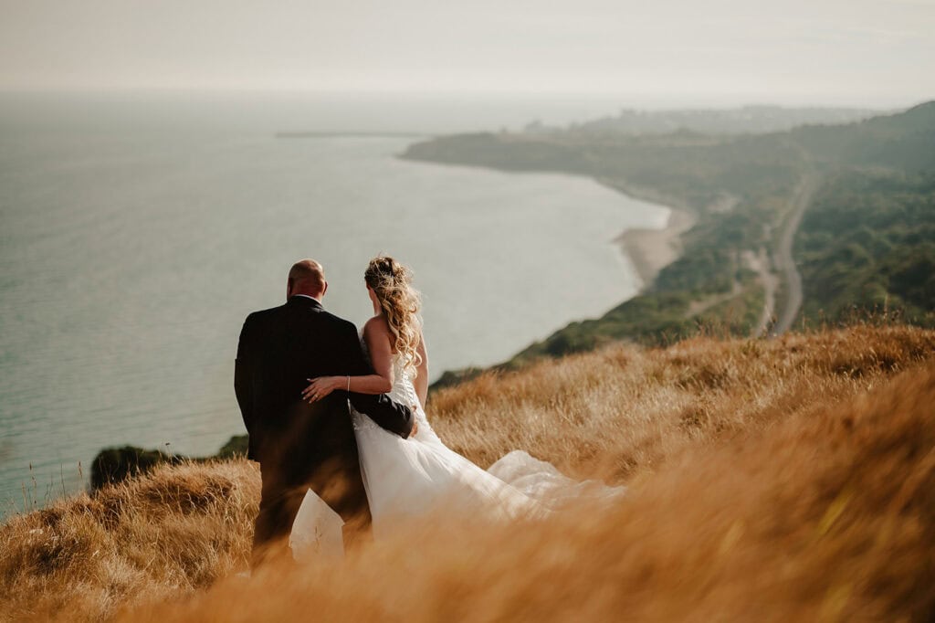 A couple wearing wedding attire, with the bride in a white dress and the groom in a black suit, sits on a grassy hillside overlooking a vast body of water and a distant coastline on a sunny day. Photographed by Dover Wedding Photographer, the scene is serene and romantic, capturing their special moment. Image by Pearce Wedding Photography.