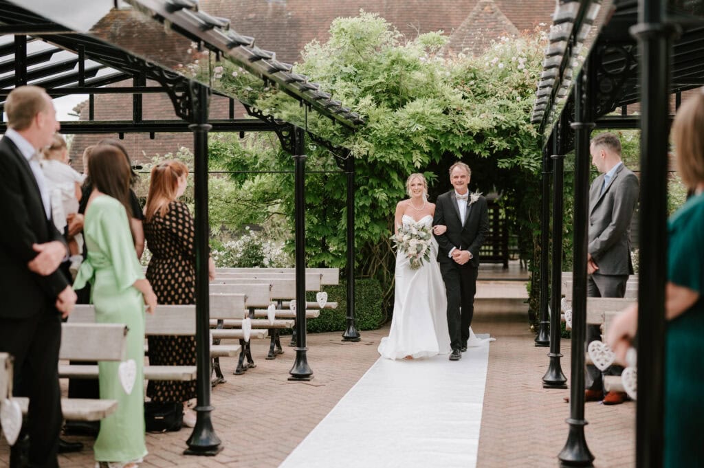 A bride in a white dress and veil walks arm in arm with an older gentleman down an outdoor aisle lined with guests on either side. The aisle, covered with a white runner, leads to a pergola set against lush greenery at The Old Kent Barn, perfectly captured by a Dover wedding photographer. Image by Pearce Wedding Photography.