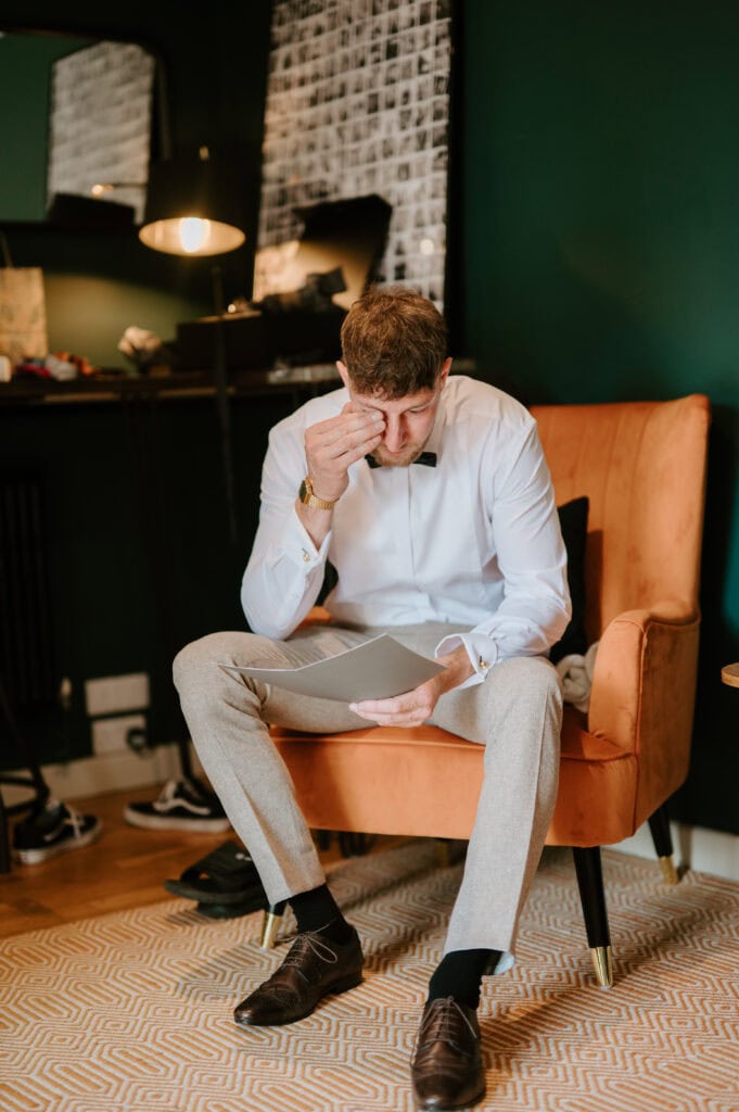 A man in a crisp white shirt and bow tie sits on an orange chair, looking stressed as he holds a piece of paper—a scene captured with the keen eye of a Kent wedding photographer. He covers his eyes with one hand; behind him, a dark wall, patterned rug, and dimly lit lamp set the mood. Image by Pearce Wedding Photography.
