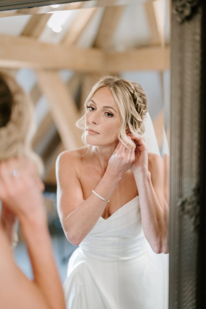 A woman in a white strapless dress adjusts an earring while looking in a mirror, captured through the lens of a Kent wedding photographer. She has blonde hair styled in an updo, reflected against the elegant background with visible wooden beams. Image by Pearce Wedding Photography.