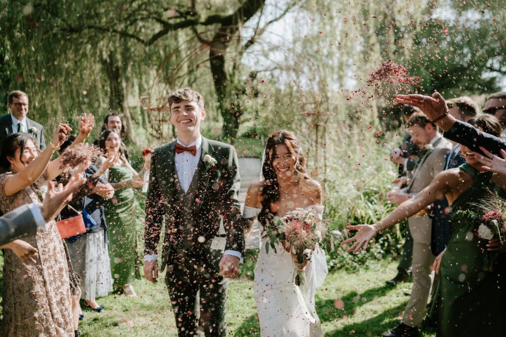 A newlywed couple walks joyfully down an outdoor aisle in Happy Valley, Norfolk, surrounded by guests throwing flower petals. The groom is dressed in a dark suit with a red bow tie, and the bride wears a white dress, holding a bouquet. Trees and greenery create a lush backdrop for their wedding day. Image by Pearce Wedding Photography.