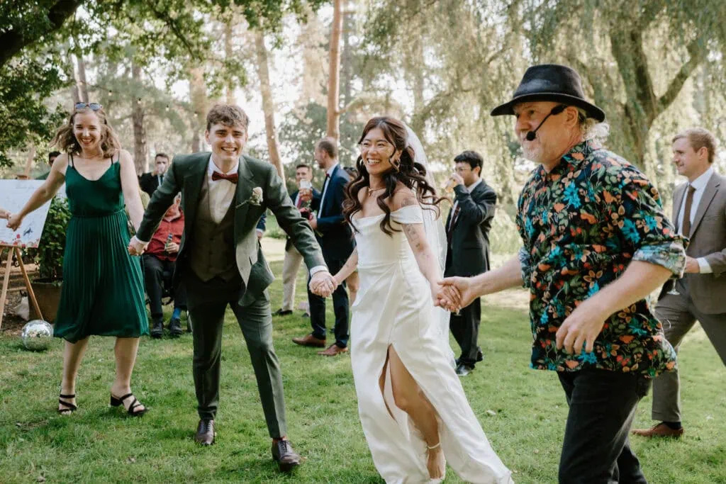 A joyous wedding celebration outdoors in Happy Valley, featuring a bride in a white dress and groom in a green suit holding hands and dancing with guests. One guest wears a floral shirt and hat. The group is smiling and surrounded by lush Norfolk greenery and trees. Image by Pearce Wedding Photography.