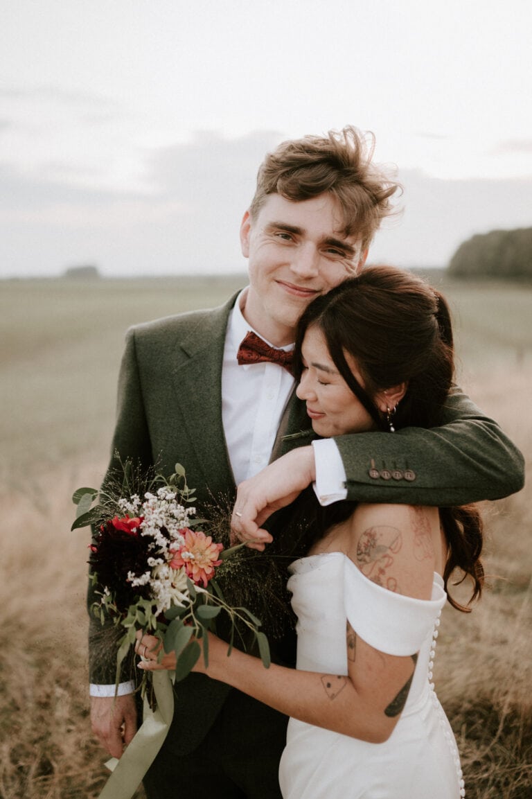 A couple stands in a grassy field during sunset at Happy Valley, Norfolk. The groom, in a green suit and red bow tie, has his arm around the bride, who is in an off-shoulder white dress. She holds a bouquet of flowers and leans into his chest with her eyes closed and a content smile on their wedding day. Image by Pearce Wedding Photography.