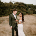 In the heart of Norfolk's Happy Valley, a bride and groom stand in a field. The bride, in an off-the-shoulder white dress holding a bouquet of flowers, and the groom, in a green suit with a red bow tie, gaze at each other smiling. Trees stand tall in the background, framing their love-filled wedding day. Image by Pearce Wedding Photography.