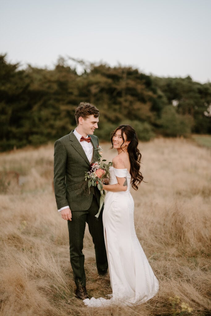 In the heart of Norfolk's Happy Valley, a bride and groom stand in a field. The bride, in an off-the-shoulder white dress holding a bouquet of flowers, and the groom, in a green suit with a red bow tie, gaze at each other smiling. Trees stand tall in the background, framing their love-filled wedding day. Image by Pearce Wedding Photography.