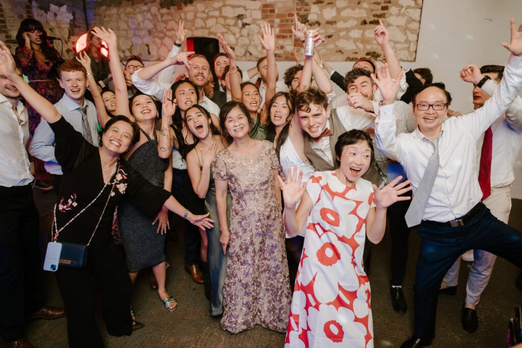 A large group of people, dressed in formal and semi-formal attire, stand together indoors. Many are smiling and raising their hands in a celebratory manner. The background includes a brick wall, adding charm to this Norfolk wedding at Happy Valley. It appears to be a joyful gathering or party. Image by Pearce Wedding Photography.