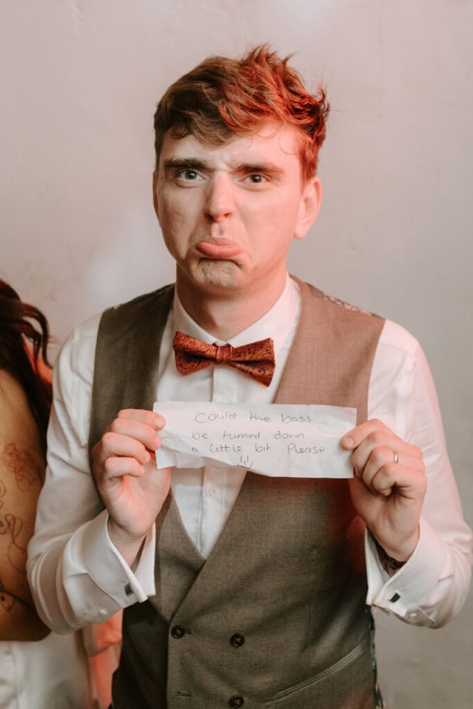 A person with a sad expression, wearing a white shirt, brown bow tie, and grey vest, holds up a handwritten note. The note reads, "Could the bass be turned down a little bit please." The scene appears to be from a wedding in Happy Valley, Norfolk. The background is a plain wall. Image by Pearce Wedding Photography.