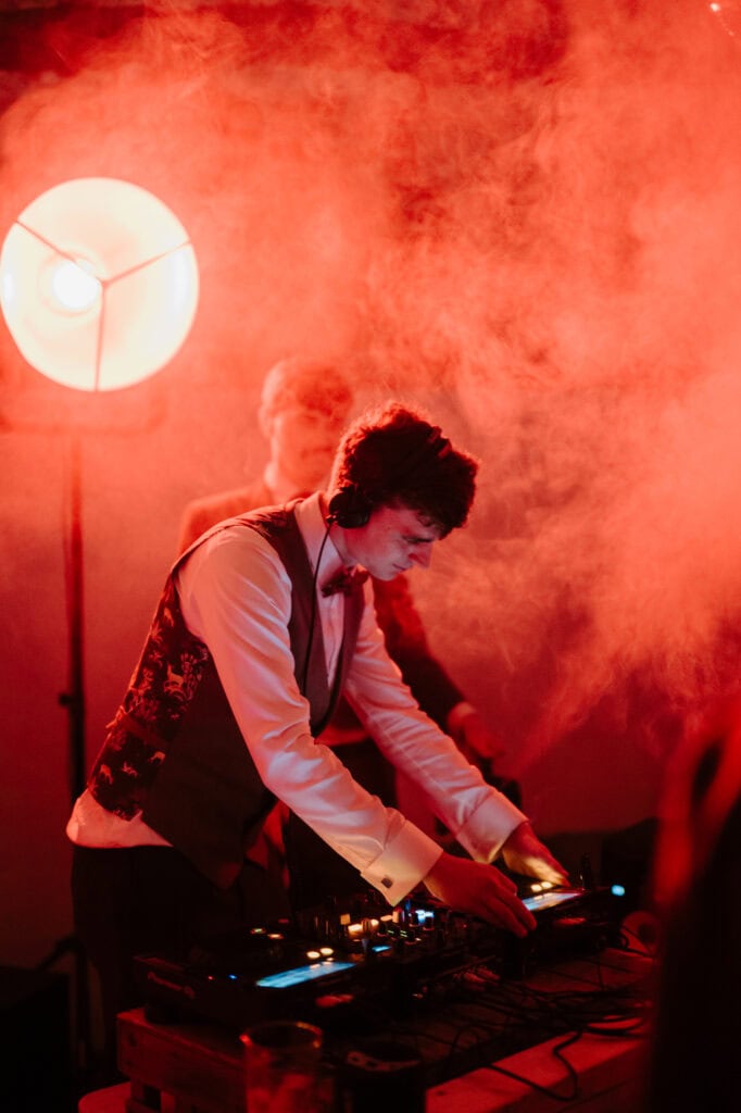 A groom wearing a white shirt and vest is DJing at a club or party, perhaps in Happy Valley or Norfolk. They are focused on the DJ equipment, with a red light and smoke creating an atmospheric effect around them. Another person is blurred in the background. Image by Pearce Wedding Photography.