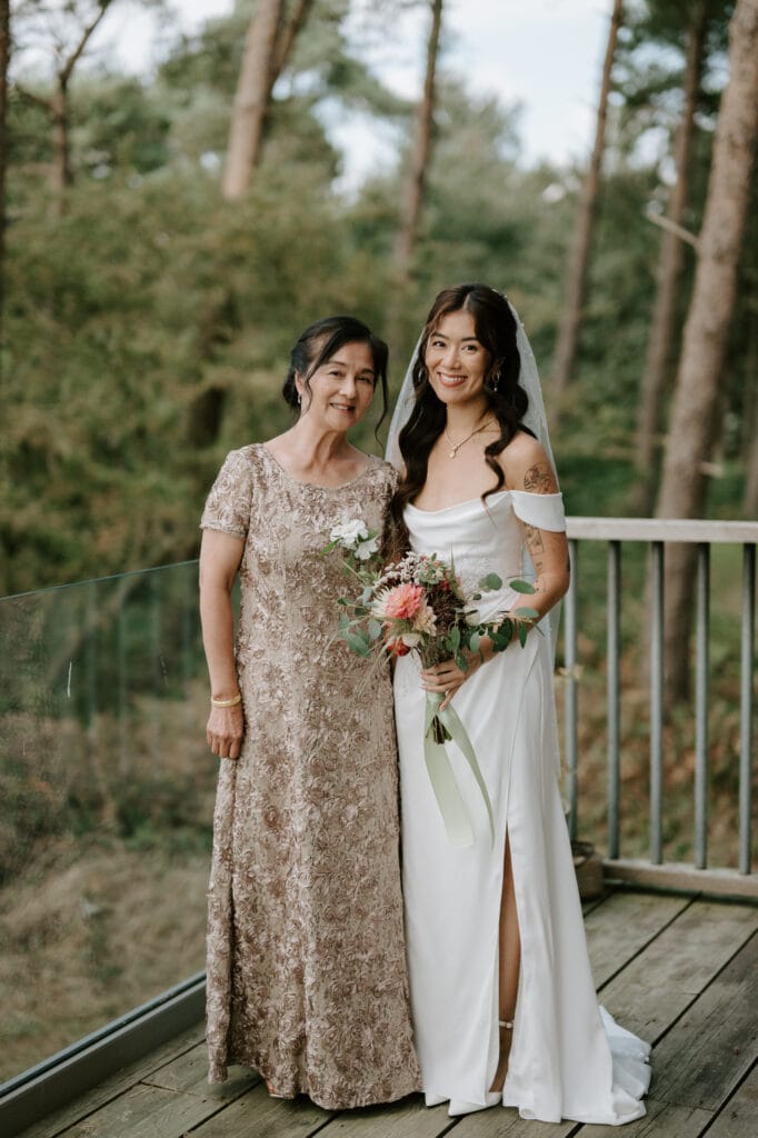 A bride in a white wedding dress and veil stands beside her mum in a beige lace dress on a wooden deck with a glass railing. Both are smiling, and the bride is holding a bouquet of flowers. Tall trees form the background, creating a serene setting that feels like Happy Valley. Image by Pearce Wedding Photography.