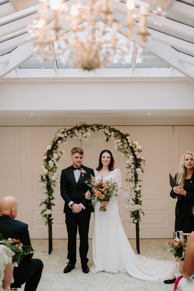 A bride and groom stand before an arch adorned with white flowers during their wedding ceremony at Hayne House in Kent. The bride is wearing a long white dress and holding a bouquet, while the groom is in a black suit with a bow tie. Guests are seated on either side, and a chandelier hangs above. Image by Pearce Wedding Photography.