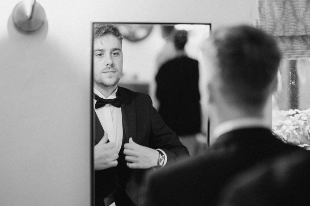 A man in a tuxedo adjusts his bow tie while looking into a mirror, preparing for a wedding at Hayne House. He appears focused and is standing in a well-lit room. The background reflection shows a blurred view of another person and interior elements. The photo is in black and white. Image by Pearce Wedding Photography.