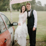 A bride and groom stand beside a red car on a rainy day at Hayne House in Kent. The smiling bride, in a white lace gown, holds a white umbrella with red hearts. The groom, in a black vest and bow tie, smiles at the camera. They are outdoors with a grassy background, capturing their perfect wedding moment. Image by Pearce Wedding Photography.