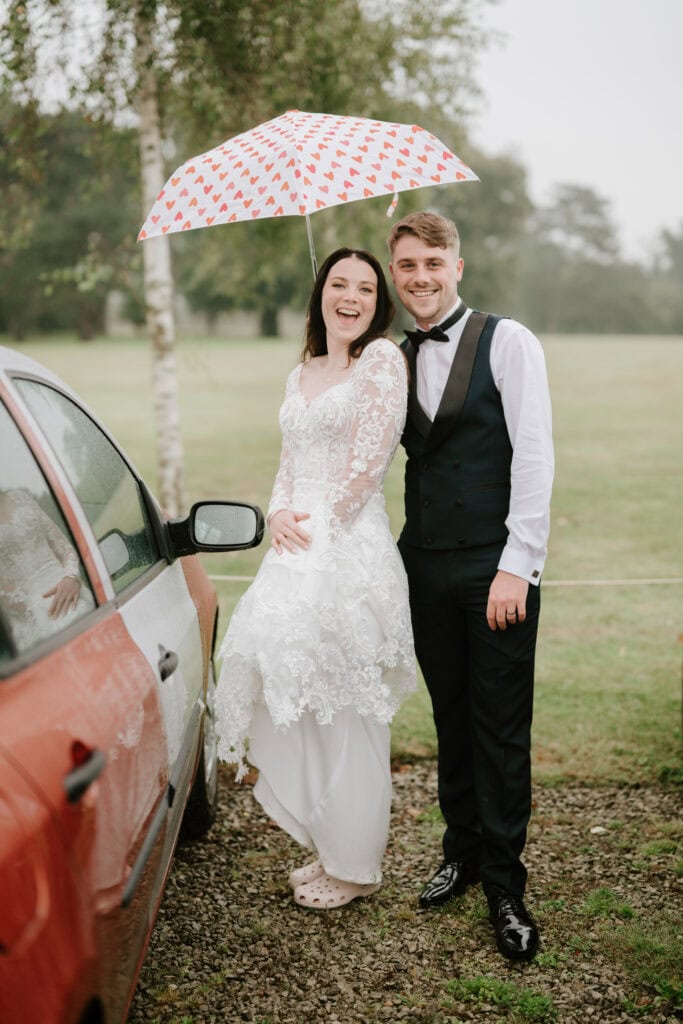 A bride and groom stand beside a red car on a rainy day at Hayne House in Kent. The smiling bride, in a white lace gown, holds a white umbrella with red hearts. The groom, in a black vest and bow tie, smiles at the camera. They are outdoors with a grassy background, capturing their perfect wedding moment. Image by Pearce Wedding Photography.
