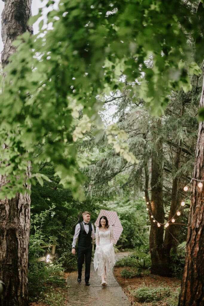 A couple walks down a forested path holding hands at their wedding in Kent. The groom wears a vest and tie, while the bride, with her white dress and pink umbrella, complements the string lights hung on the lush green trees. Light rain falls gently on this magical scene near Hayne House. Image by Pearce Wedding Photography.