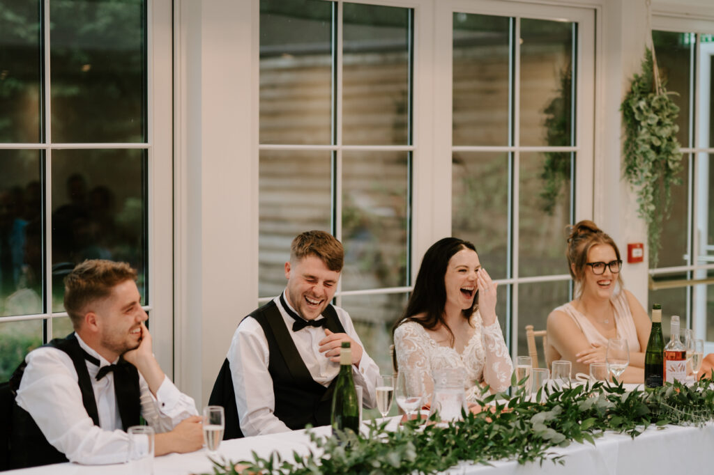 Four people, dressed formally, sit at a long table adorned with greenery decoration. They are all laughing and appear to be at a wedding reception at Hayne House in Kent. Behind them are large glass doors. Various drinks, including wine, are on the table. Image by Pearce Wedding Photography.
